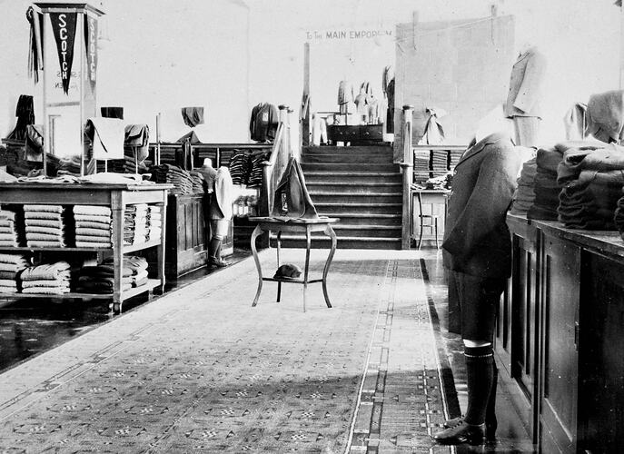 Section of clothing shop with boyswear. Tables of folded clothes at left and a mannequin in school uniform on right. Central patterned walkway.