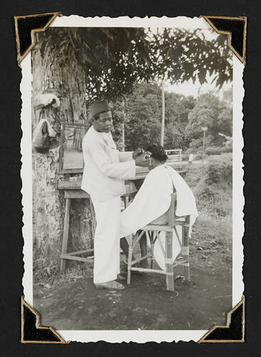One standing man cutting the hair of a seated man in front of tree.