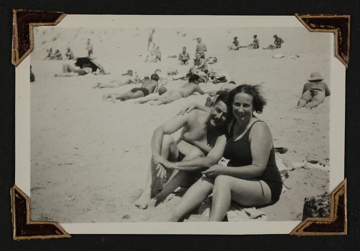 Man and woman in bathers sitting in beach, people sunbathing in background.
