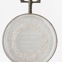 Round medal with text framed by wreath, text around. Suspension loop at top.