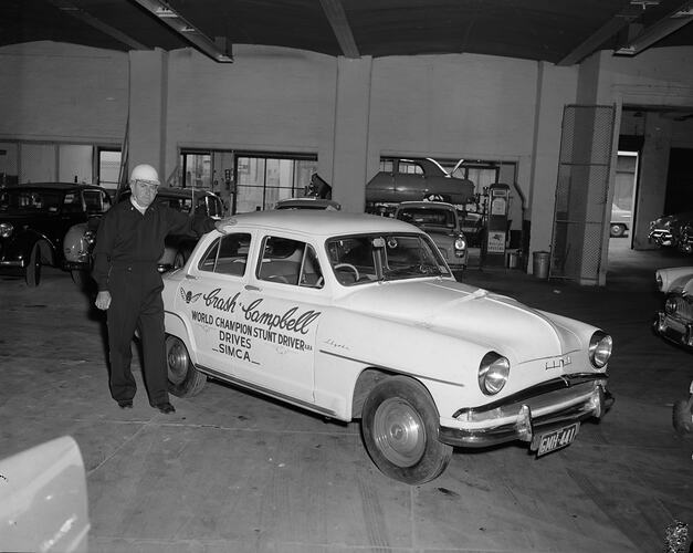 Stuntcar driver wearing crash helmet standing next to motor car. Motor vehicles and a Mobilgas pump in background.