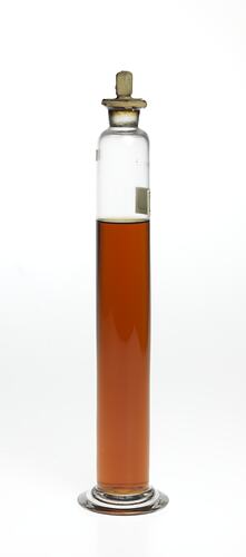 Cylindrical glass jar with amber coloured liquid. Two labels affixed, sealed at top.