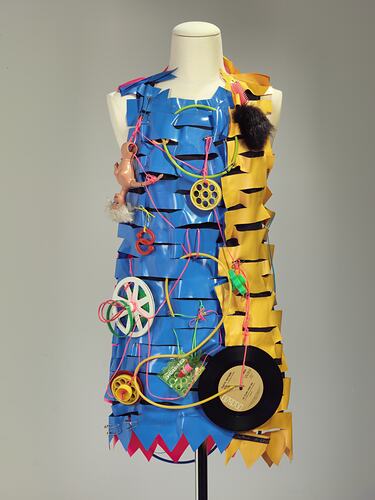 Dress made of horizontal strips of pink, yellow, blue plastic. Adorned with plastic cables, dolls, objects.