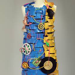 Dress made of horizontal strips of pink, yellow, blue plastic. Adorned with plastic cables, dolls, objects.