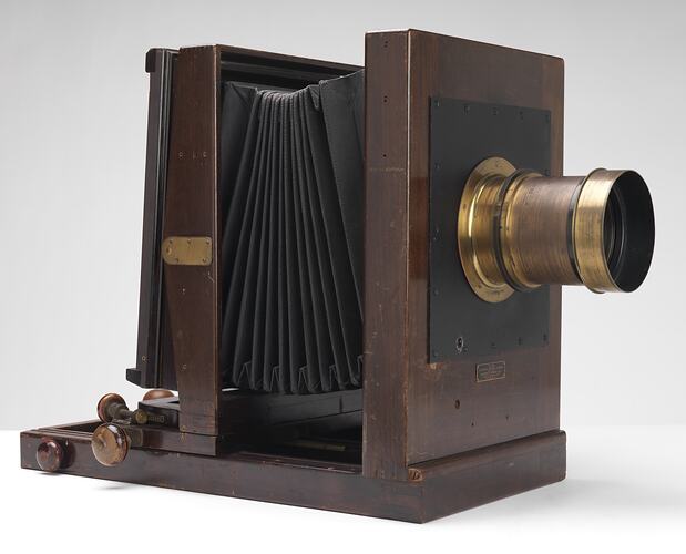 Dark wooden box-shaped camera with central cylindrical brass lens.