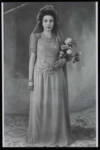 Woman in wedding dress and headpiece holding flowers in her left hand. Studio setting.