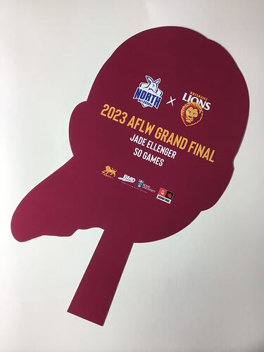 Back of cut-out card face. Maroon with yellow and white printing. Handle at base.