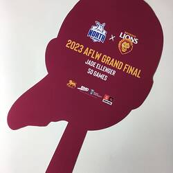 Back of cut-out card face. Maroon with yellow and white printing. Handle at base.