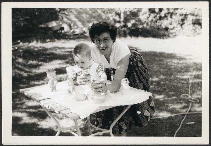 Woman holds baby seated on wooden table in outdoor garden setting.