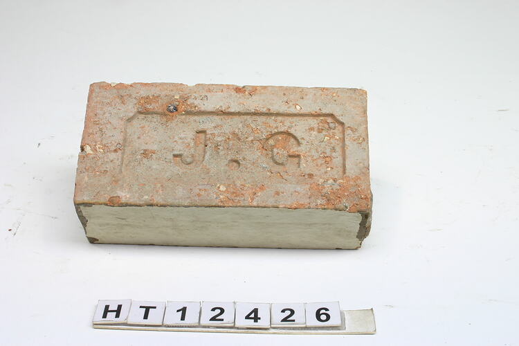 Red rectangular machine pressed brick lightly covered with remnant cement. Impressed letters "J.G.".