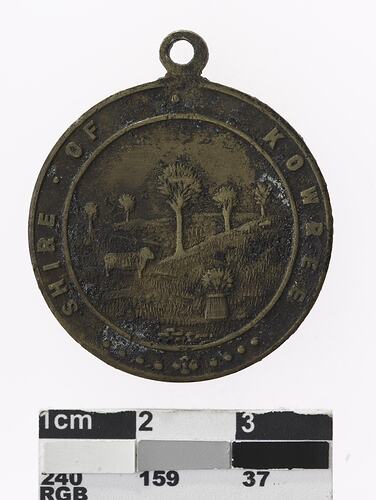 Round medal with pastural scene featuring tress and a sheep, text surrounding.