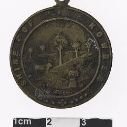 Round medal with pastural scene featuring tress and a sheep, text surrounding.