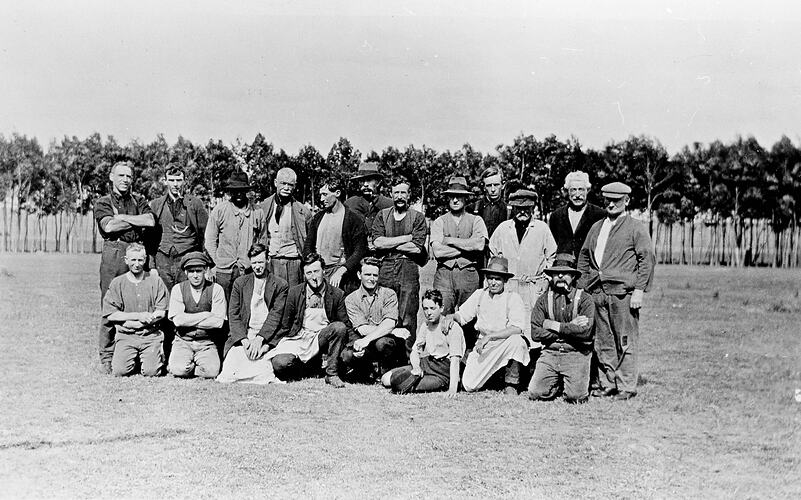 Group portrait of twelve men standing behind eight men and boys sitting on the ground outside.