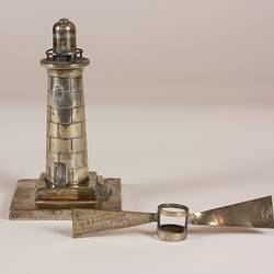 Silver lighthouse paperweight. Beside it, a removed part, silver blade-like light beams with a central ring.