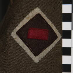 Felt patch with white diamond, topped with brown diamond, topped with maroon rectangle sewn onto green fabric.