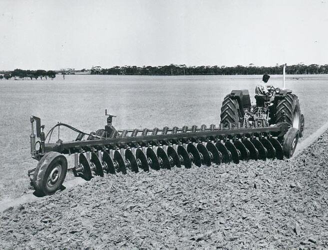 Rear view of a plough being drawn by a man driving a tractor across a wide open field.