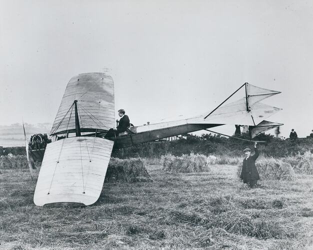 Man sitting in monoplane in corn field and man holding tail.