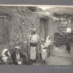 Village men, women and children in a narrow street. It is lined with drystone and mud walled buildings with th