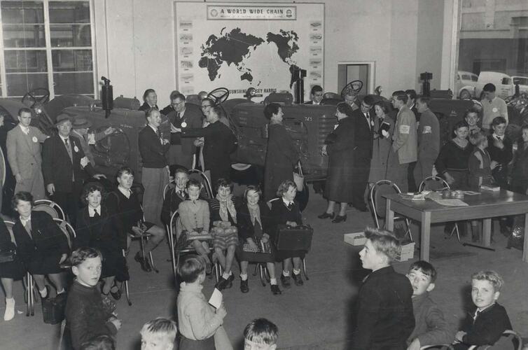 Crowd of people and seated school children in showroom.