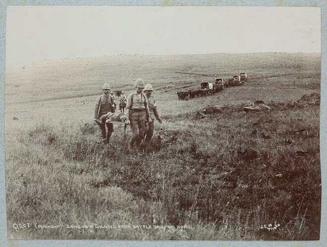 Three soldiers carrying man on stretcher, convoy of wagons and horses In background.