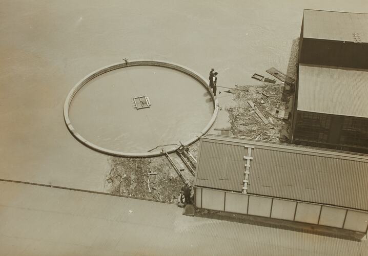 Aerial view of flooded river. A tank is full of water with two men standing on the rim looking at the water.