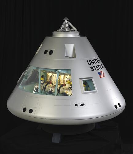 Conical shaped silver spacecraft model. Three seated astronauts inside facing upwards.