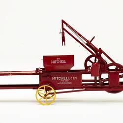 Red metal hay press model with turning gears and springs, four yellow wheels and white painted letters on side
