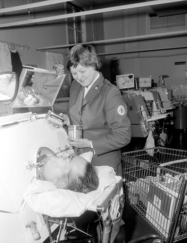 Woman holds up soap and tinned fruited to a man laying in an iron lung. A shopping trolley is beside her.