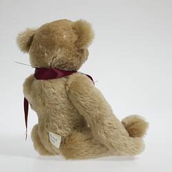 Back of seated light brown plush bear with red ribbon around neck.
