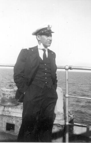 Man wearing suit and captain's hat stands on a ship deck. Ocean in the background.