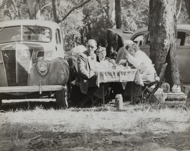Men and women eating a picnic at a table next to a 1937 Ford sedan in bushland.