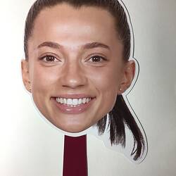 Cut-out card face of female smiling. She has a brown pony tail. Handle at base.