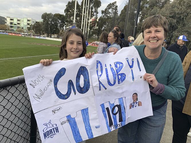 Girl and woman holding a white paper handmade banner with blue letters. Football oval behind them.