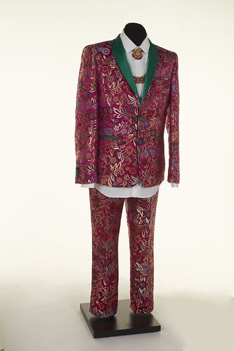 Pink suit jacket and pants with ornate pattern and green lapels. White collared shirt and ornate necklace.