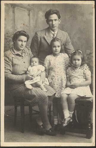 Family portrait of five. Man stands at back whilst a woman and two girls are seated. The woman holds as baby.