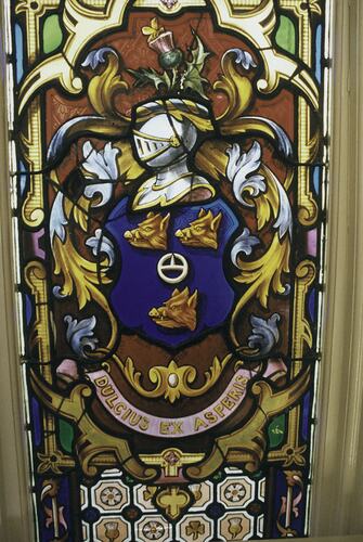Stained glass window showing a coat of arms. Metal helmet with visor above 3 boars. Scroll below.