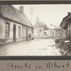 House lined street with one building bomb damaged and damaged carts on right side of road.