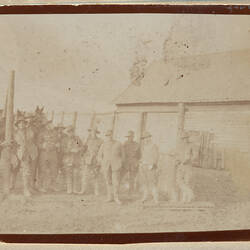 Group of servicemen with horsed behind them, standing in front of a wooden house.