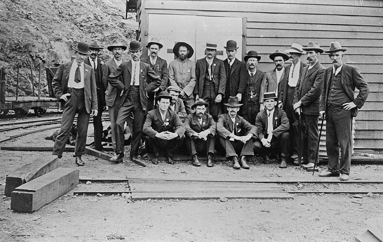Group of men outside a building of the Mt Morgan Mine. They are all dressed in suits and are wearing hats and ties.