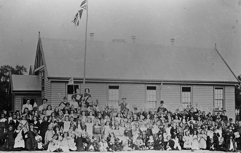 Pupils and teachers assembled outside wooden school building. Flagpole has a Union Jack flag.