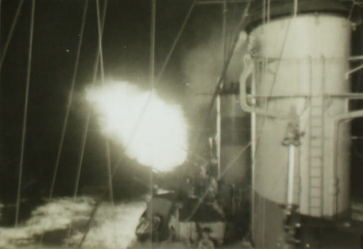 Military battleship with explosion in the air to left of ship, ocean in the left background.