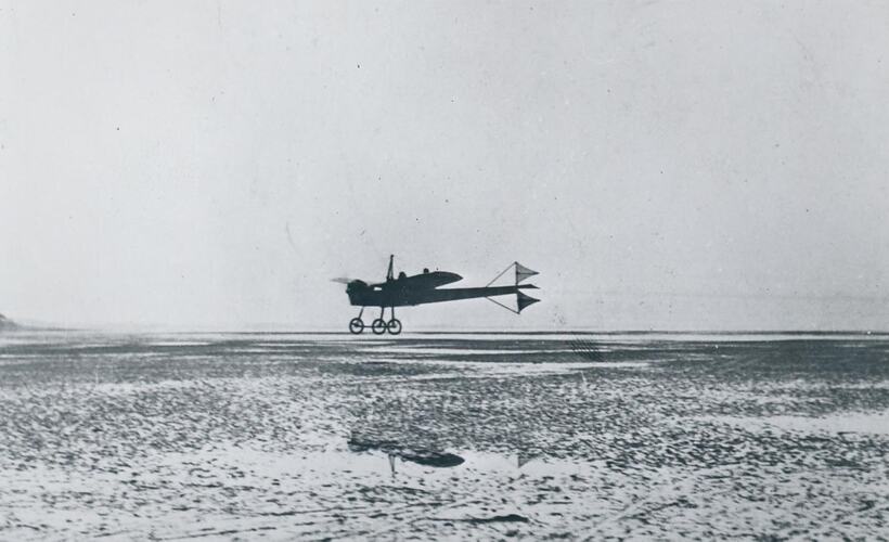 Monoplane flying over wet sand.