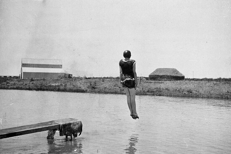 Girl wearing bathing suit and cap doing a pin drop, feet first off a jetty into a dam.