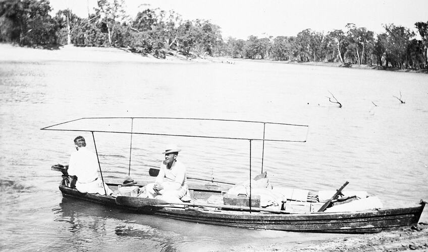 Two men in a boat drawn up by the riverbank. The boat has framework for an awning.