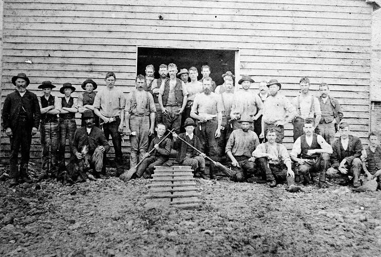 Shearers out front of a shearing shed. Two boys with crossed brooms in centre foreground.