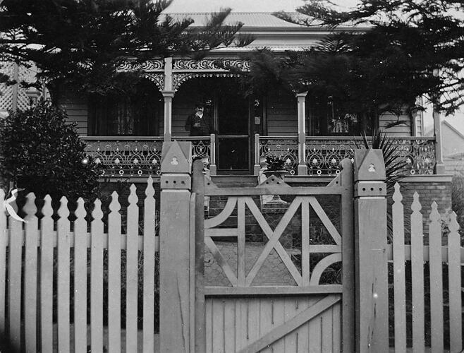 Man standing on verandah of a small wooden house with an elaborate iron lacework verandah. Picket fence and gate in foreground.