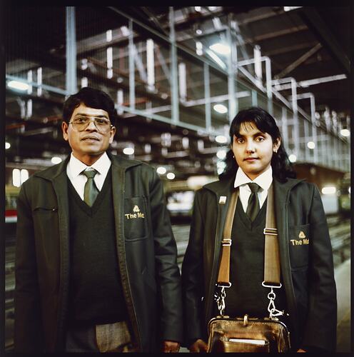 Man and woman stand in tram depot wearing green uniforms. Woman has a brown tram ticket satchel.