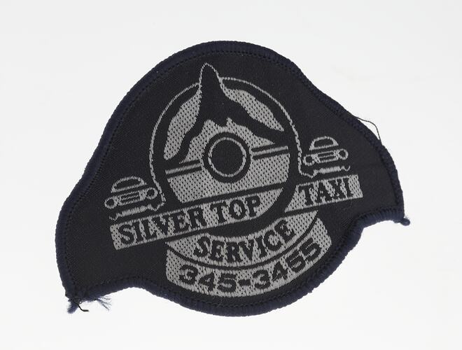 Dark cloth badge with white embroidered lettering below two cars framing a mountain top over a steering wheel.
