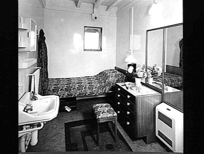 Ship interior. Single bed against wall. Handbasin at left, chest of drawers and heater at right.