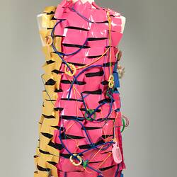 Back of dress made of horizontal strips of pink, yellow, blue plastic. Adorned with plastic cables, dolls, obj